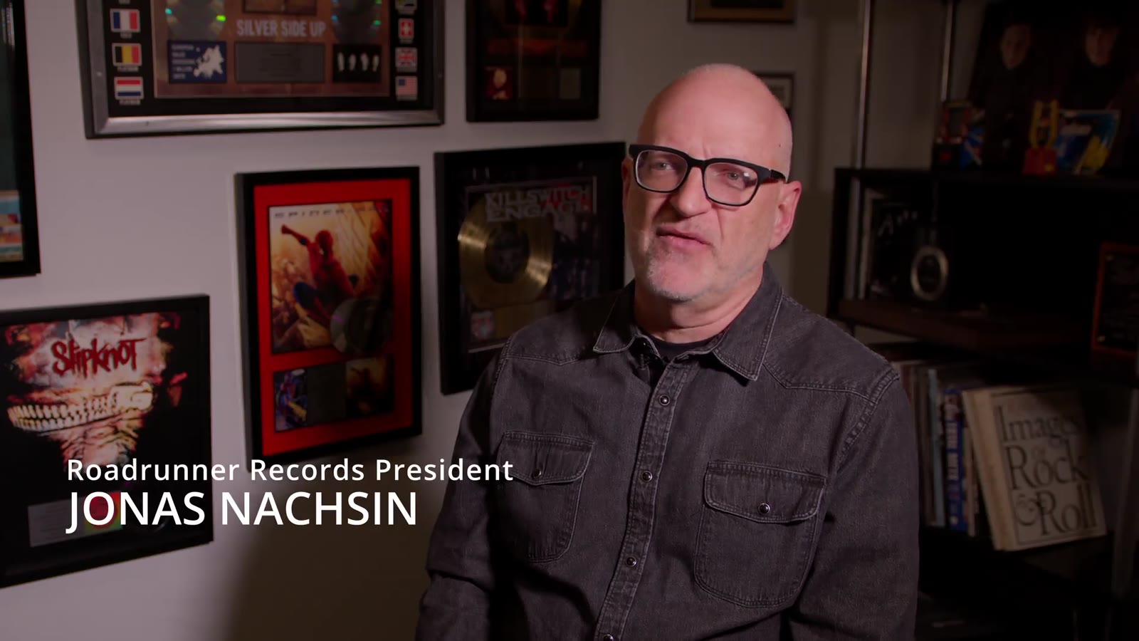 A record-label executive being interviewed on camera in a room hung with framed album art